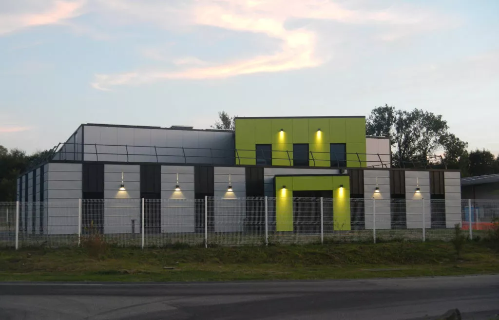 Façade du bâtiment du Groupe H2RA au crépuscule, architecture moderne gris et vert avec éclairages extérieurs allumés derrière une clôture.