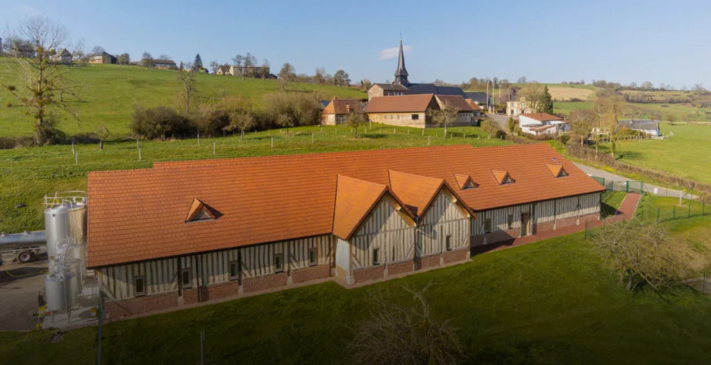 Vue aérienne d’une usine de camembert à Camembert, long bâtiment à colombages avec grande toiture en tuiles terre cuite, entouré de prairies et d’un village au loin, chantier de couverture en Normandie par l’entreprise H2RA.